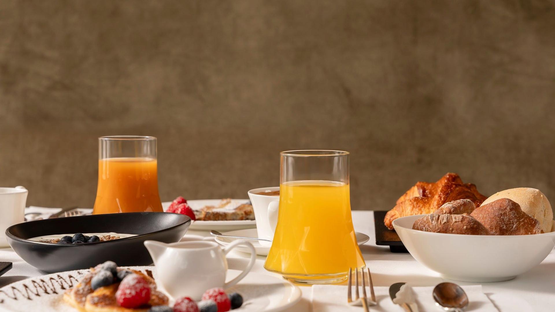 Colazione con succo d'arancia, pane, croissant e frutta fresca su tavola imbandita.