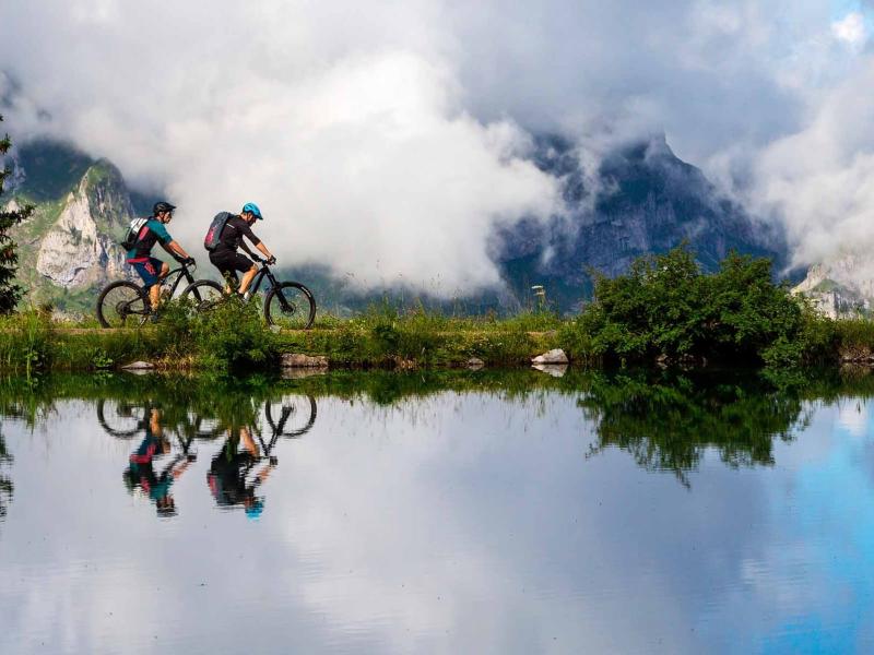 Ciclisti su un sentiero montano, riflessi nell'acqua, con montagne e nuvole sullo sfondo.