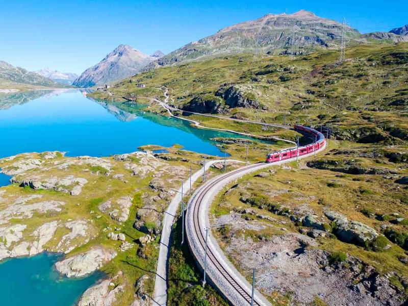Treno rosso attraversa paesaggio montano con lago e montagne riflessi nell'acqua cristallina.