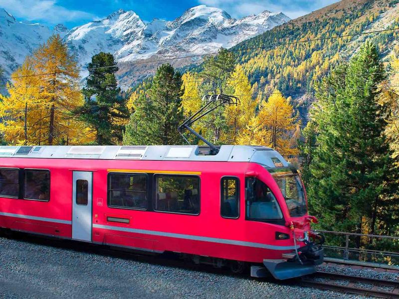 Treno rosso attraversa foresta autunnale con montagne innevate sullo sfondo.