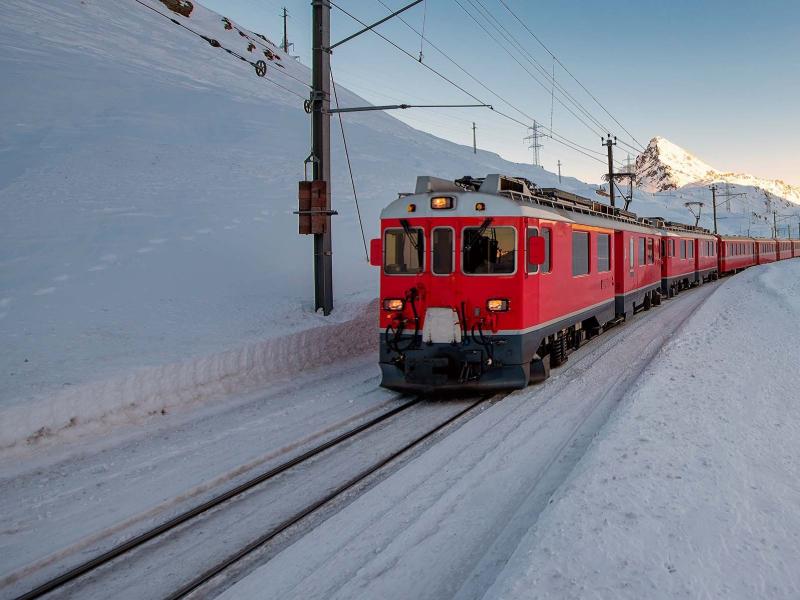 Treno rosso attraversa paesaggio innevato al tramonto, montagne sullo sfondo.